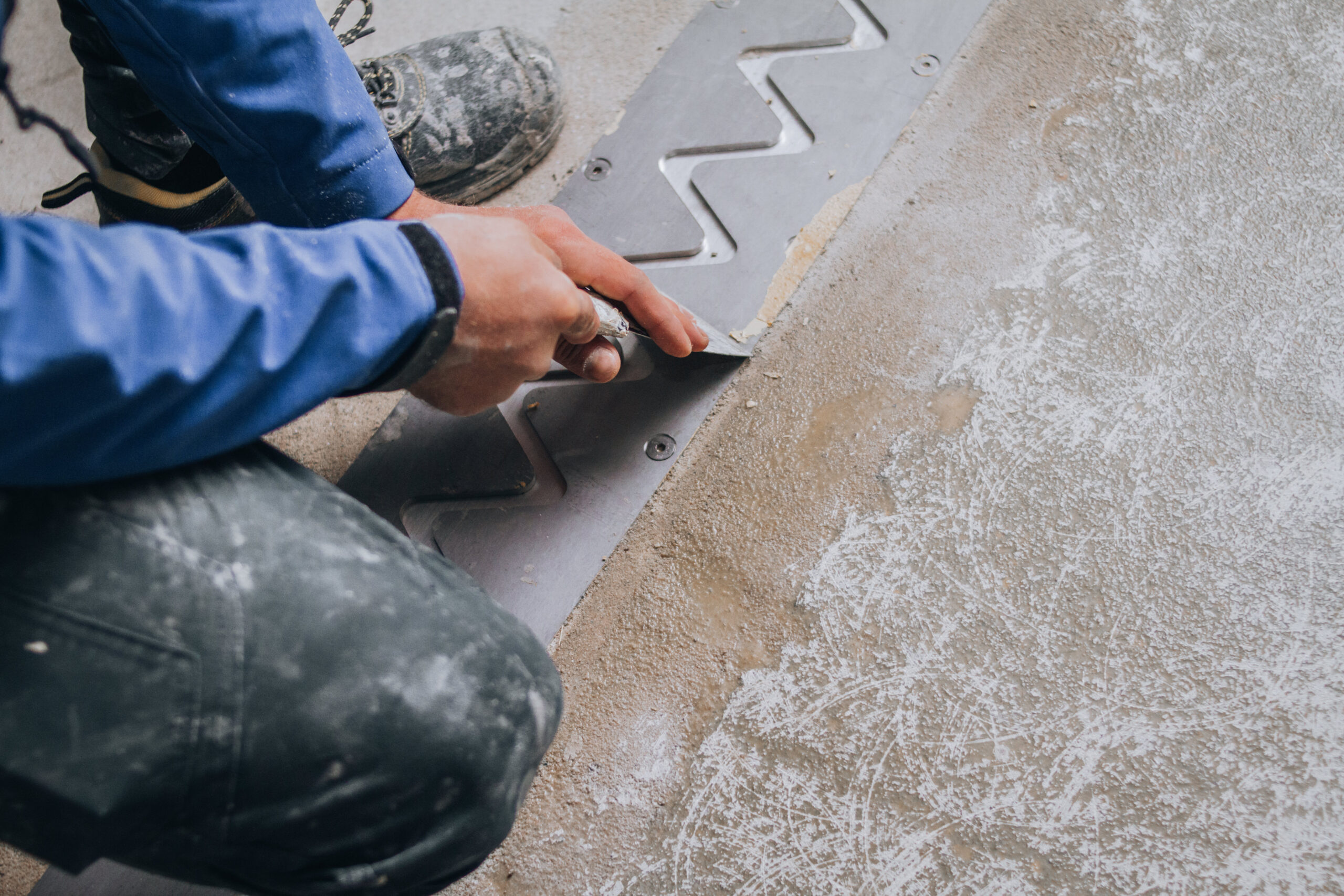 worker's skilled hands installing floor expansion joints. The worker is shown wearing protective gear and kneeling down on the concrete floor, using specialized tools to carefully measure and cut the material to fit precisely.